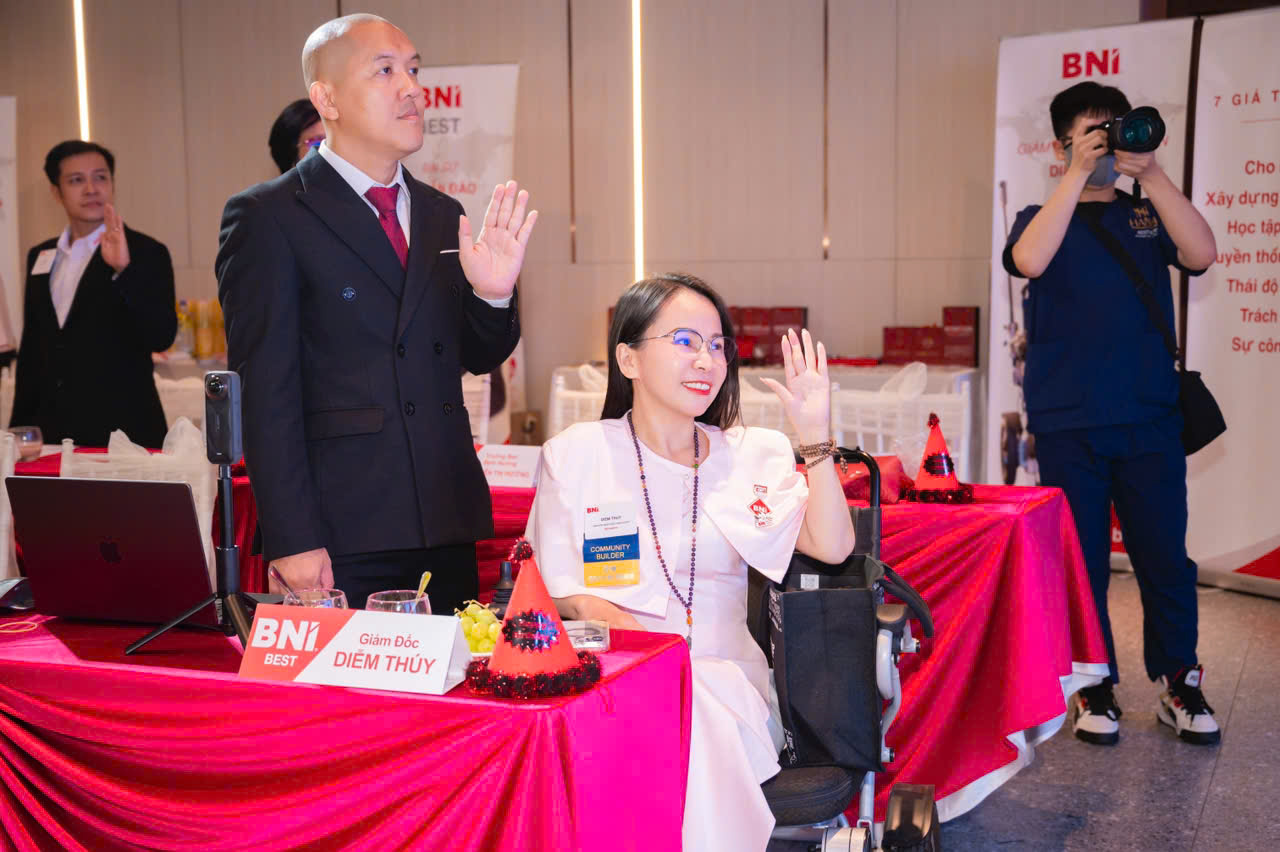 Coach Diễm Thúy giơ tay vẫy chào bên cạnh một lãnh đạo nam, bàn phủ khăn đỏ BNI