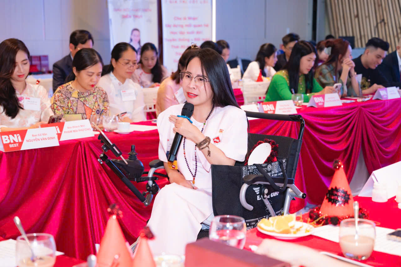 Coach Diễm Thúy cầm micro chia sẻ giữa hội trường, mọi người ngồi quanh bàn phủ khăn đỏ