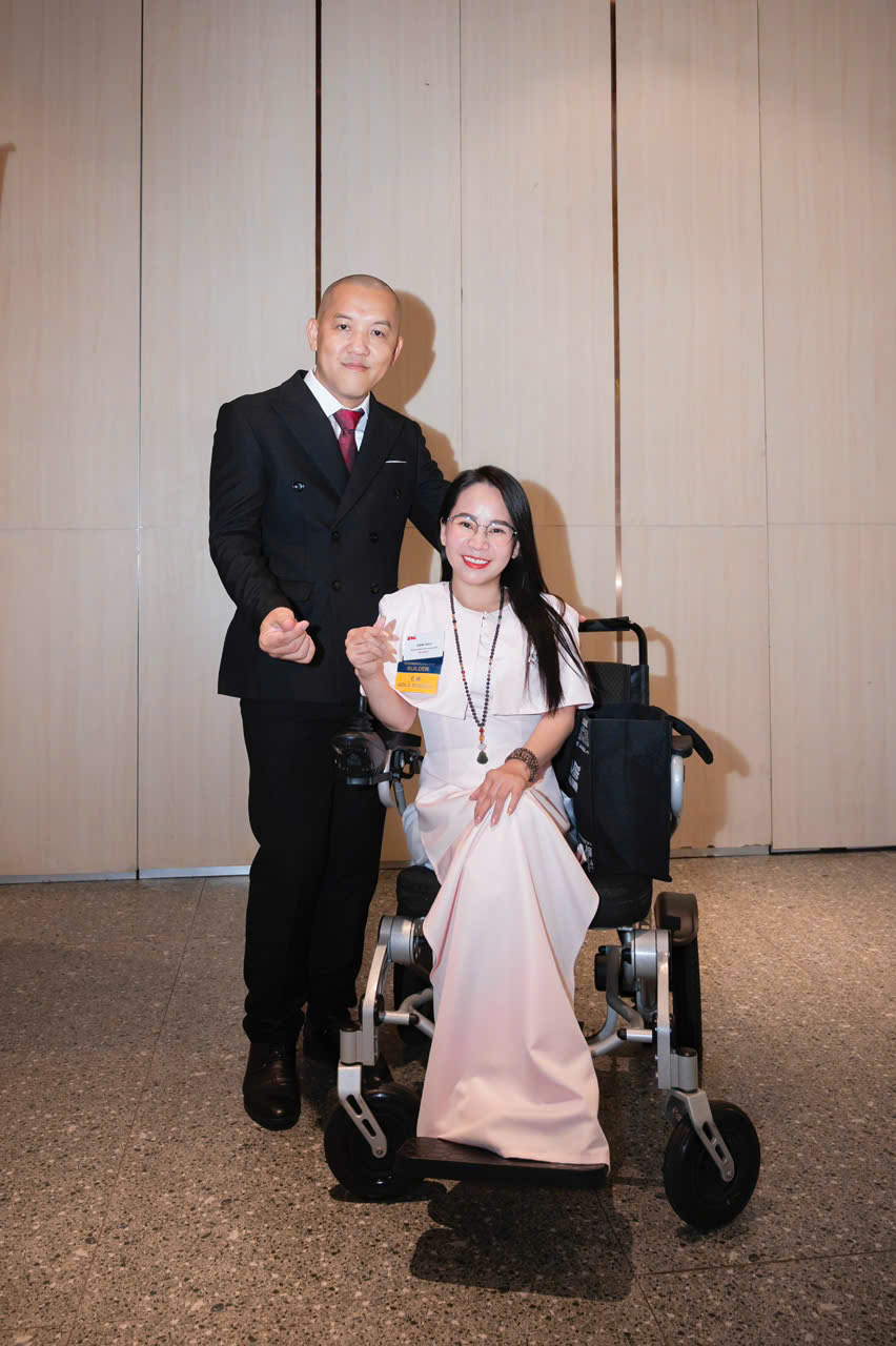 Coach Diễm Thúy ngồi xe lăn, mặc áo hồng nhạt, bên cạnh một lãnh đạo